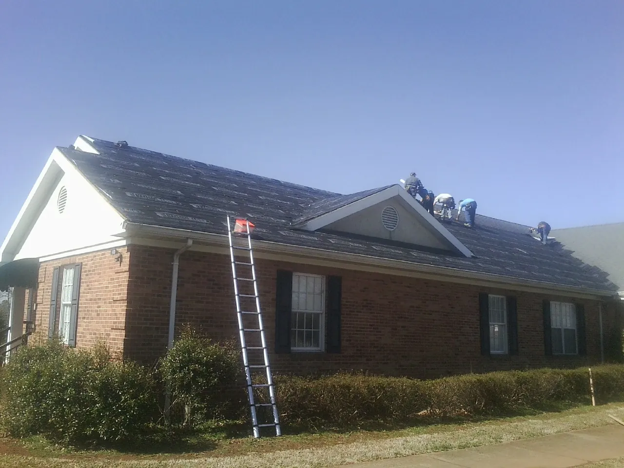 Crew tearing shingles off a brick ranch roof with a ladder set — Metal Roof Repair work by our crew in Sheldahl