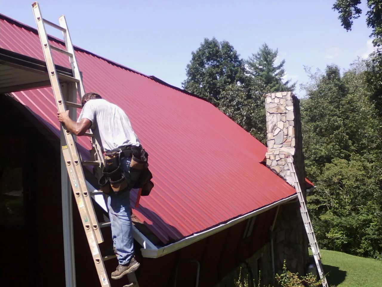 Roofer on a ladder beside a red metal roof and stone chimney — Soffit & Fascia Repair by Roofing Brothers in Sheldahl